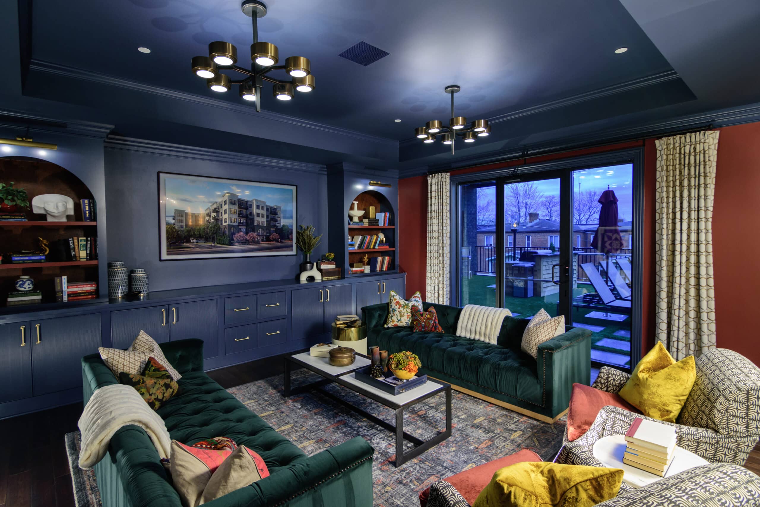 Modern living room with blue ceiling and cabinets, green velvet sofas, bookshelves, large wall art, coffee table, and sliding glass doors overlooking an outdoor patio at dusk.