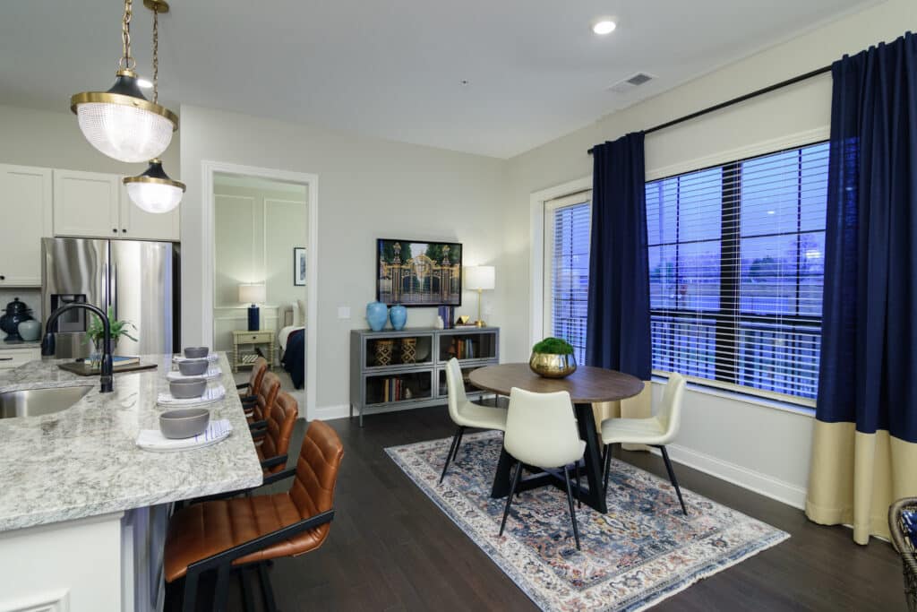 Modern open-plan kitchen and dining area with marble countertops, leather barstools, round dining table, four chairs, large window, and a TV on a console beside a rug.