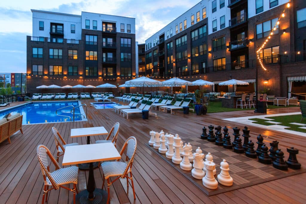 Outdoor lounge area with a swimming pool, lounge chairs, white umbrellas, string lights, and a large chess set on a wooden deck in front of an apartment building at dusk.
