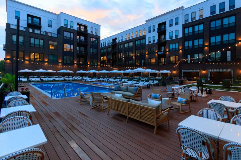 Outdoor pool area at an apartment complex with lounge chairs, patio tables, string lights, and surrounding modern buildings at sunset.