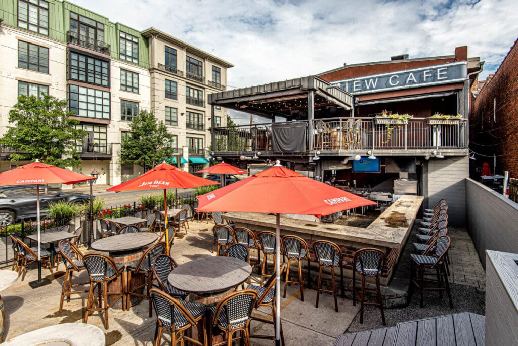 Outdoor cafe patio with round tables and red umbrellas, adjacent to a two-story building with a sign reading "CAFE" and surrounded by modern apartment buildings.