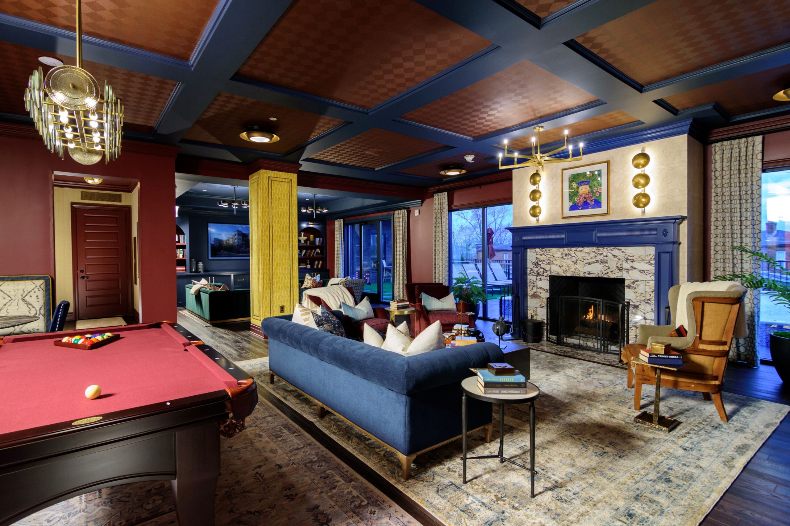 A stylish living room with a blue sofa, armchairs, fireplace, pool table, bookshelves, and large windows revealing an outdoor view.