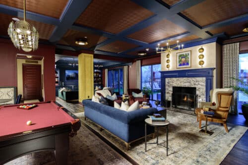 A stylish living room with a pool table, blue sofa, armchair, fireplace, bookshelves, and large windows showing an outdoor view.