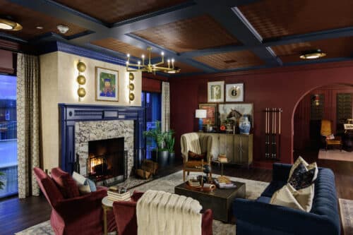 A living room with a lit fireplace, red and navy walls, armchairs, a sofa, a coffee table, decorative art, and a chandelier, featuring warm, ambient lighting.