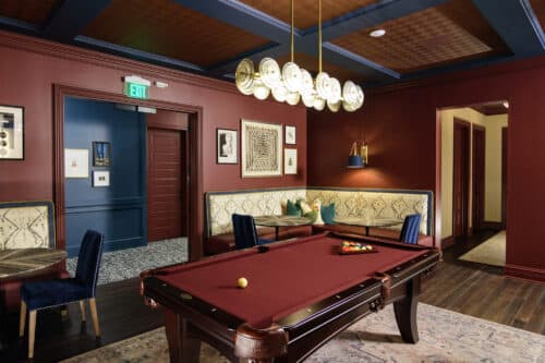 A room with a red pool table, built-in cushioned seating, wall art, round tables with chairs, and a modern chandelier overhead.