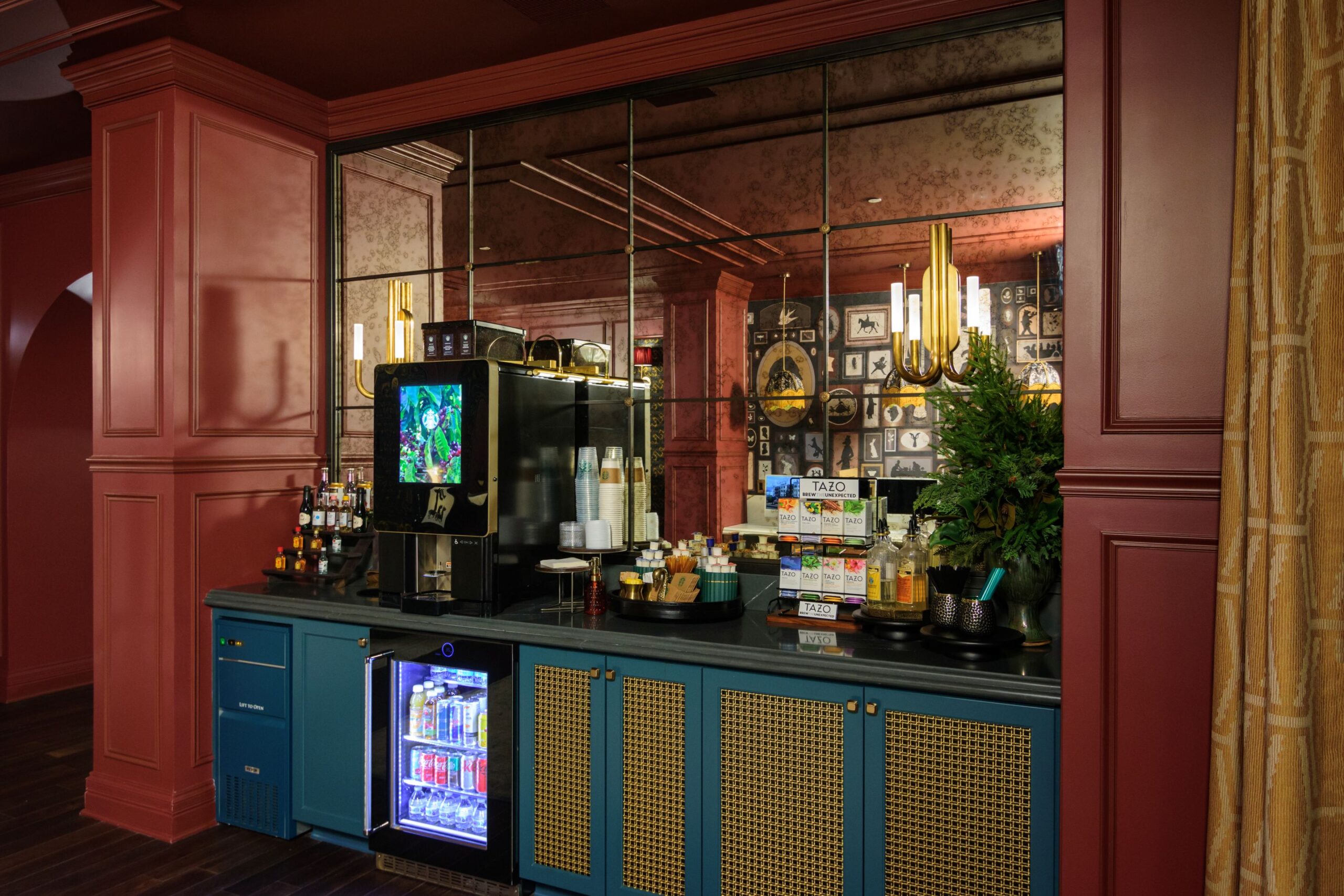 A self-serve beverage station with a coffee machine, mini fridge, assorted drinks, cups, condiments, and supplies, set against mirrored panels and red walls in a stylish room.