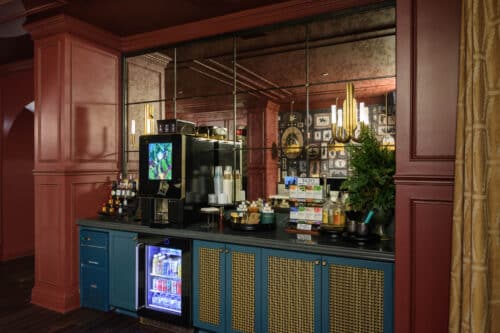 Self-serve beverage station with a coffee machine, drink fridge, condiments, cups, and various supplies, set against a mirrored wall in a room with red walls and gold accents.