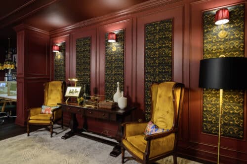 A room with burgundy paneled walls, gold-patterned insets, two yellow velvet armchairs, a dark wood desk, and red wall lamps, creating a vintage, elegant atmosphere.