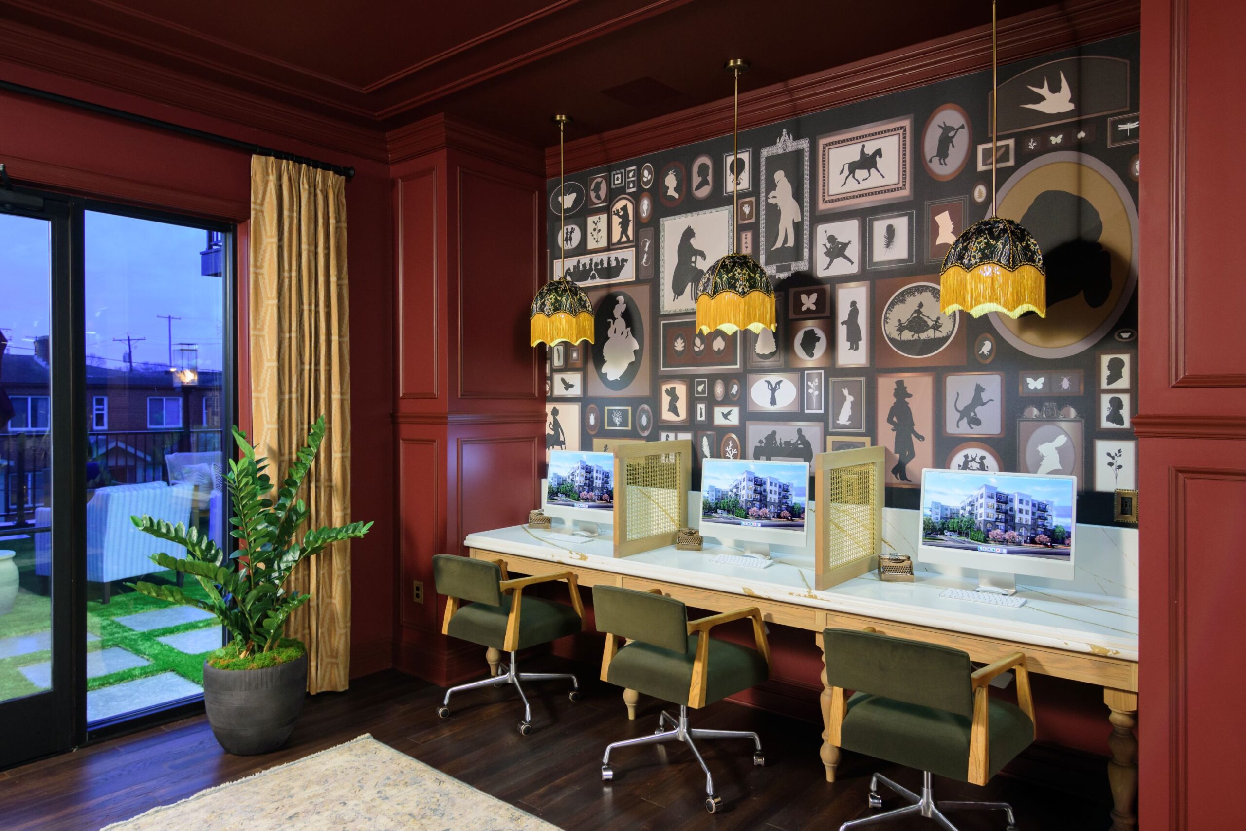 A workspace with three computers on a long desk, separated by partitions, green chairs, gold pendant lights, and a patterned wall with framed silhouettes and animal images.
