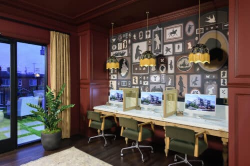 Three computer workstations with privacy screens sit against a deep red wall decorated with framed art; pendant lights hang above, and a large plant is in the corner near glass doors.