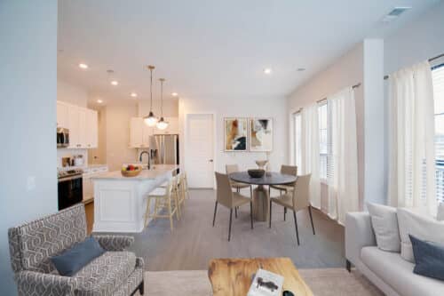 Modern open-plan kitchen and living area with white cabinets, island seating, round dining table, neutral-toned furniture, large windows, and wall art.