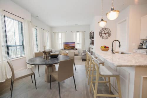 Modern open-concept living space with dining table, kitchen island with stools, and a living area featuring a sofa, TV, and large windows with curtains.