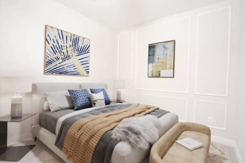 A modern bedroom with a bed, blue and beige bedding, two bedside tables with lamps, an upholstered bench, and two abstract paintings on white paneled walls.