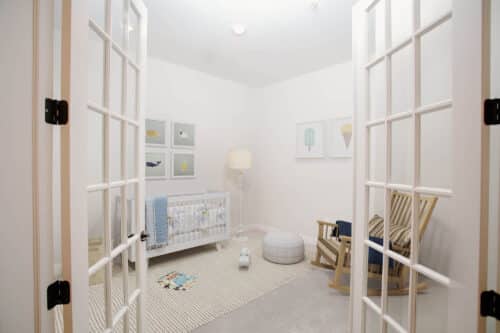 A nursery with a crib, rocking chair, pouf, lamp, and framed artwork on white walls, viewed through open French doors. Neutral and light blue accents decorate the room.