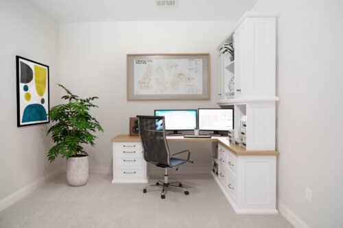 A modern home office with a desk, computer monitors, black chair, potted plant, framed art, and a city map on white walls with built-in shelves and drawers.