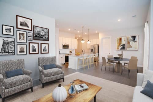 Open-concept living area with patterned armchairs, a wooden coffee table, gallery wall, dining table, and a modern white kitchen with barstools and pendant lighting.