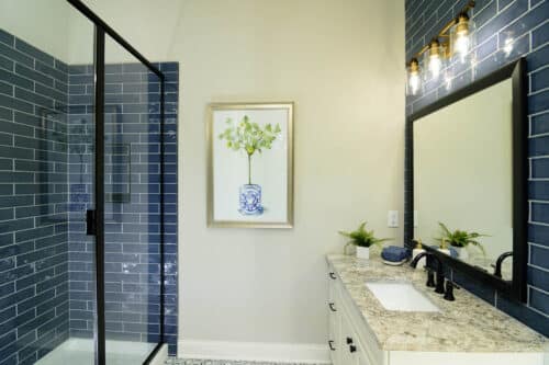 Modern bathroom with a glass shower, blue subway tiles, large mirror, and a countertop with a sink, plants, and a framed botanical artwork on the wall.