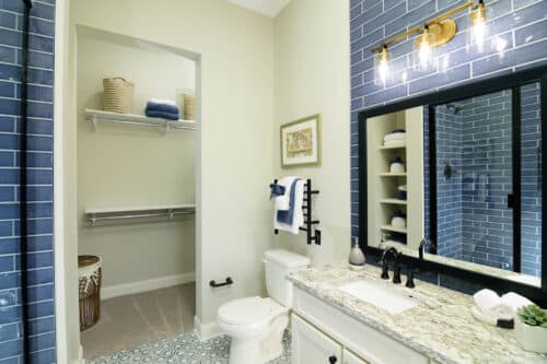 Modern bathroom with blue tiled walls, a granite countertop, white cabinetry, wall-mounted mirror, toilet, open closet with shelves, and baskets for storage.