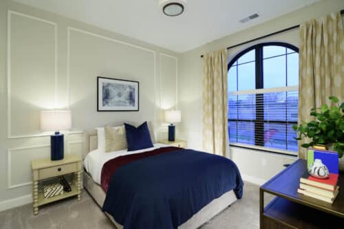 A neatly made bed with white and navy bedding stands between two nightstands with lamps; a large window with curtains and a desk with books are also visible in the bedroom.