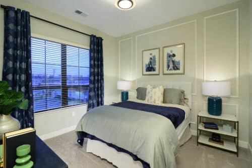 A neatly made bed with gray and navy bedding sits between two nightstands with matching lamps in a modern bedroom with framed artwork and a window with blue curtains.