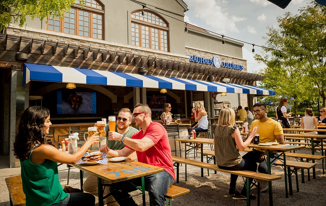 People sit at outdoor wooden tables, eating and drinking, in front of a building with a blue-and-white striped awning that says "RADHAUS OUTDOORS.