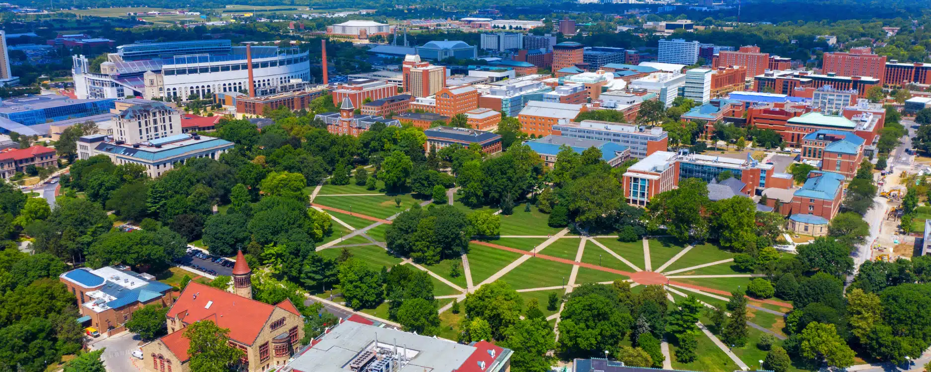 Aerial view of a large university campus with a central green quad, surrounded by academic buildings, trees, and walkways, under a clear sky.