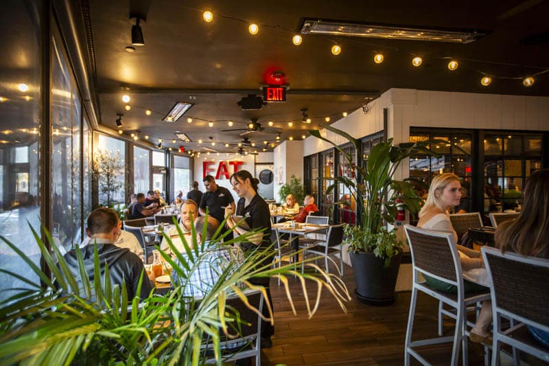 Indoor restaurant with several groups of people dining, a server taking orders, potted plants, string lights, and large windows letting in natural light.