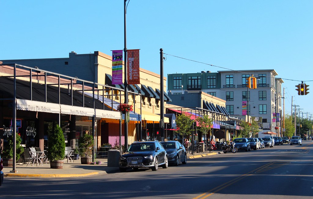 A city street with parked cars and storefronts on a sunny day; pedestrians are visible on the sidewalk near outdoor seating areas.