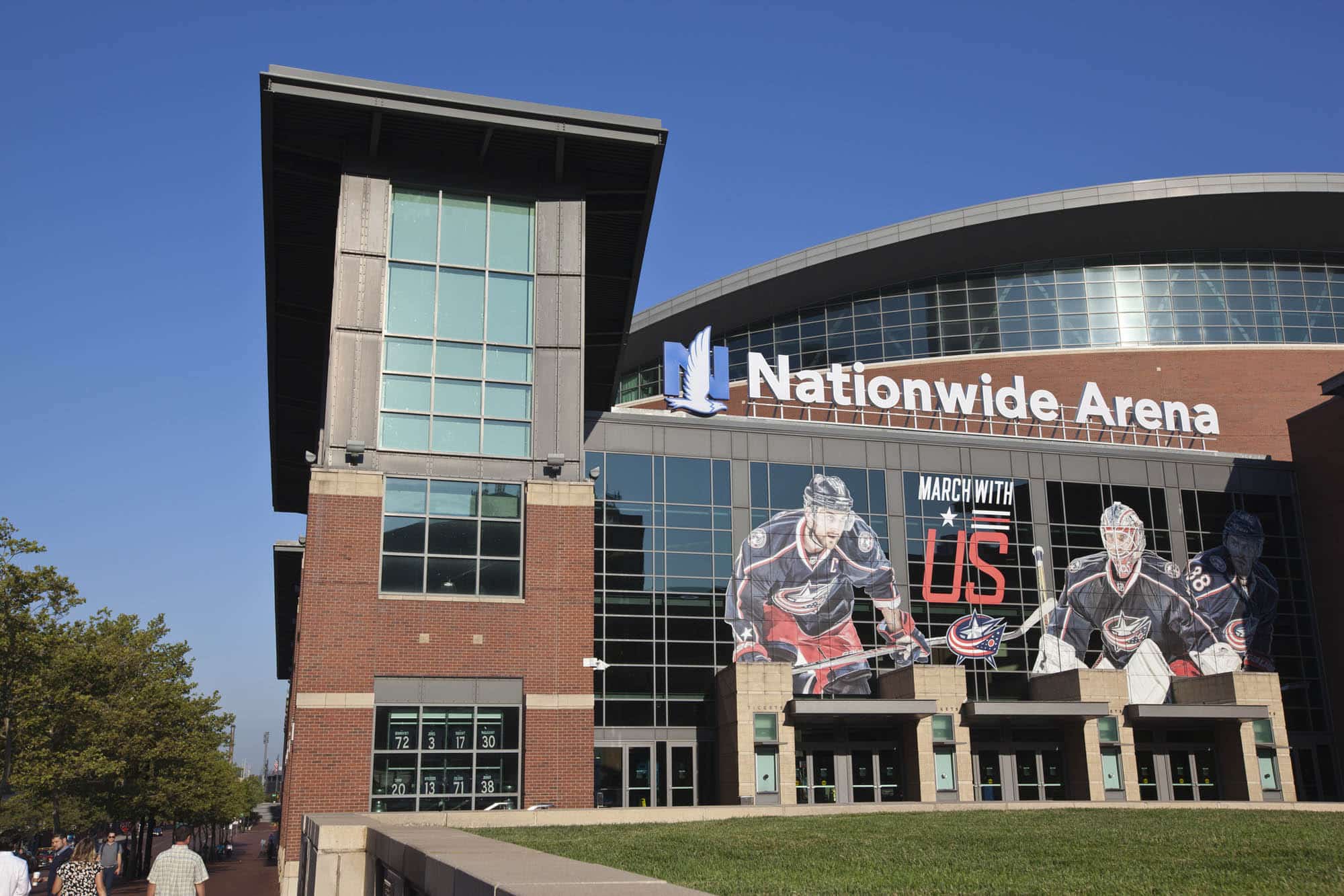 Exterior view of Nationwide Arena with large hockey player images and "March With Us" signage on glass windows, under a clear blue sky.