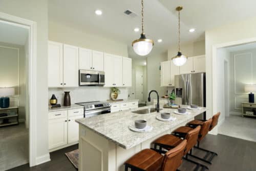Modern kitchen with white cabinets, stainless steel appliances, granite island with sink, four brown chairs, pendant lights, and place settings for four.