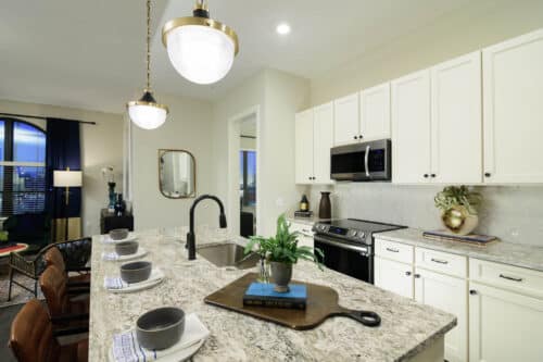 Modern kitchen with white cabinets, granite countertops, stainless steel appliances, a kitchen island with sink, and pendant lights. Place settings and a potted plant decorate the island.
