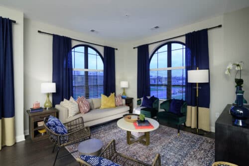 A modern living room with arched windows, navy blue curtains, a beige sofa with colorful pillows, green chairs, a rug, and various decorative items.