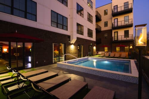 Modern apartment complex courtyard with a lit swimming pool, lounge chairs, a fire feature, and building exterior at dusk.