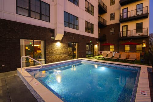 Outdoor pool at night next to a modern apartment building, with poolside loungers, red umbrellas, and windows lit from inside.