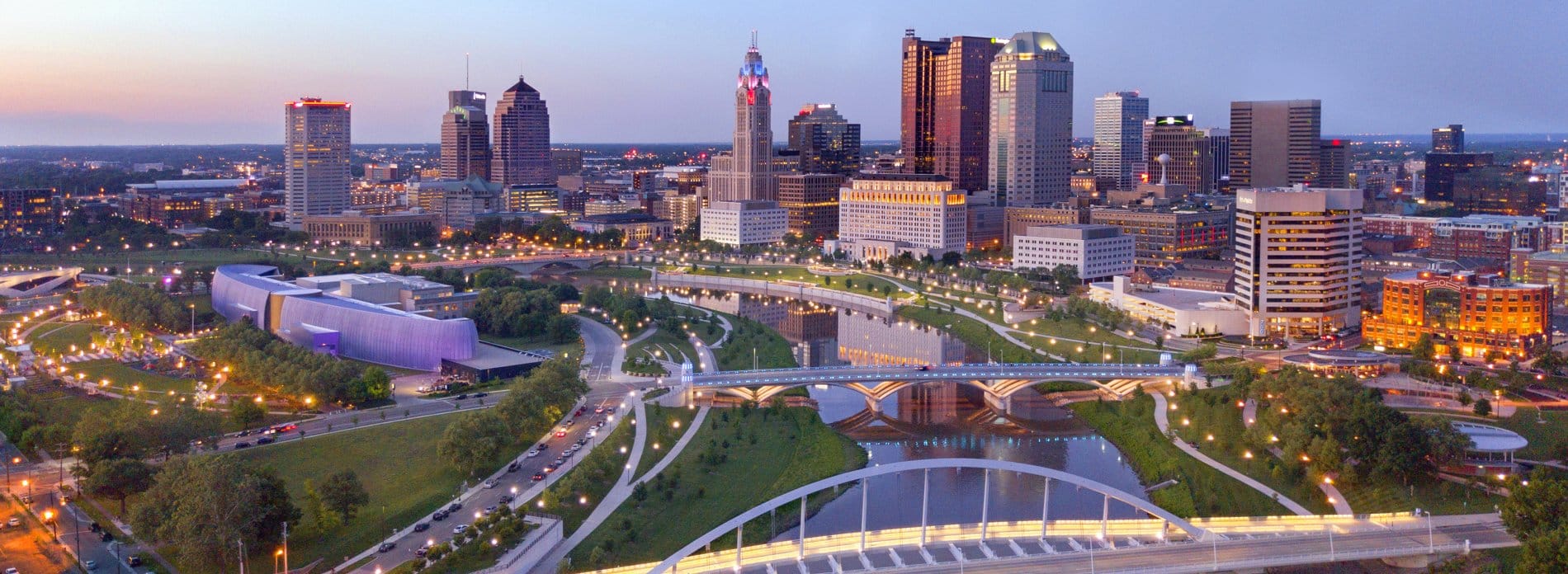 Aerial view of downtown Columbus, Ohio at dusk with high-rise buildings, bridges, and the Scioto River in the foreground.