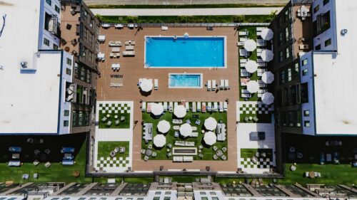 Aerial view of a rectangular swimming pool and hot tub surrounded by lounge chairs, umbrellas, and checkerboard-patterned patios in a modern apartment complex courtyard.