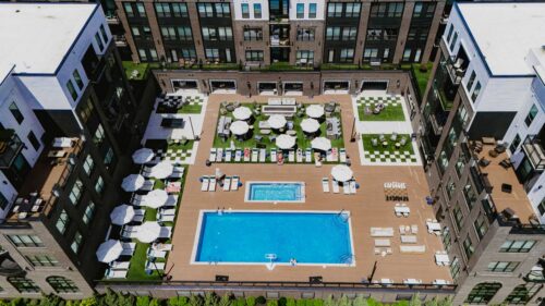 Aerial view of an apartment complex courtyard featuring a large rectangular swimming pool, lounge chairs, umbrellas, and seating areas surrounded by multi-story buildings.