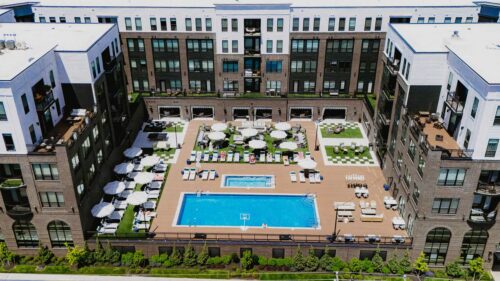 Aerial view of an apartment complex courtyard featuring a large rectangular swimming pool, lounge chairs, umbrellas, and landscaped green areas.