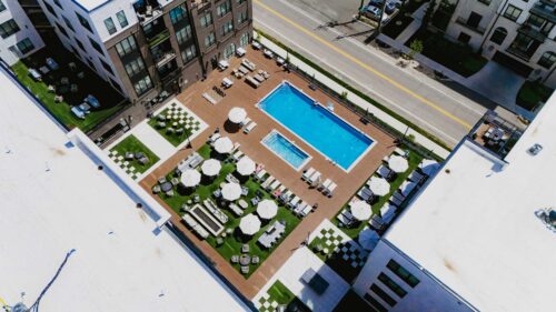 Aerial view of a modern apartment complex pool area with lounge chairs, umbrellas, greenery, and a street in the background.