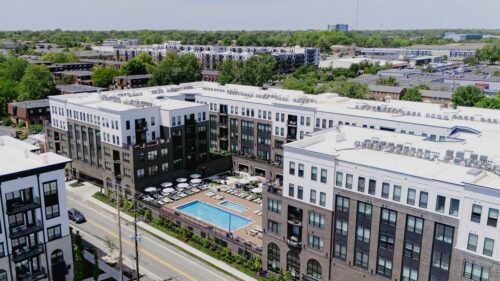 Aerial view of a modern apartment complex with a central outdoor swimming pool, surrounded by city streets, trees, and other buildings in the background.