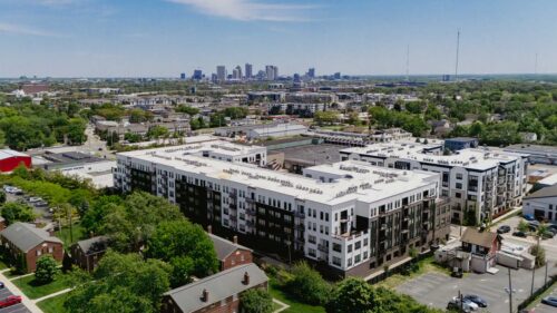 Aerial view of a modern, multi-story apartment complex surrounded by trees and neighborhood buildings, with a city skyline visible in the background under a clear sky.