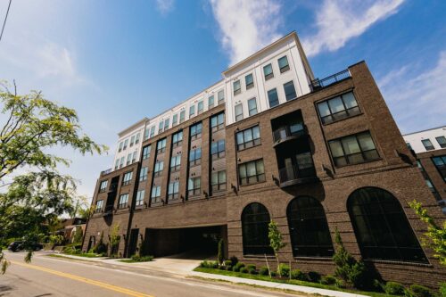 A modern multi-story apartment building with brown and white exterior walls, large windows, and balconies, situated along a quiet street under a blue sky.