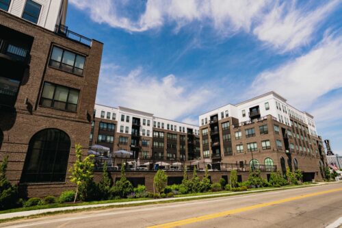Modern multi-story apartment building with dark brick and white accents, large windows, balconies, and a landscaped sidewalk along a street under a partly cloudy sky.