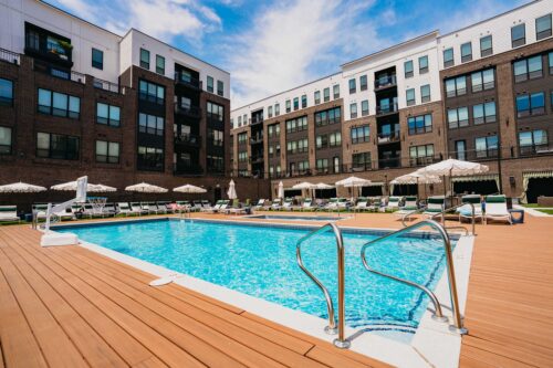 Outdoor swimming pool with metal handrails, surrounded by lounge chairs and umbrellas, set in the courtyard of a modern apartment complex under a partly cloudy sky.