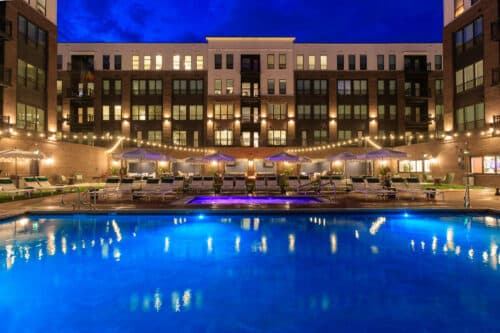 Lit apartment building with illuminated windows and balconies, string lights, umbrellas, lounge chairs, and a blue outdoor pool at night.