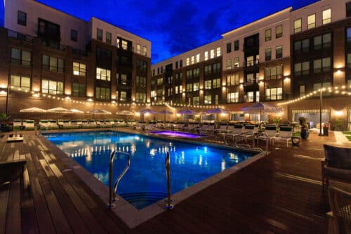 A modern apartment complex with a lit outdoor pool, lounge chairs, and string lights at night. Four-story buildings surround the pool area.