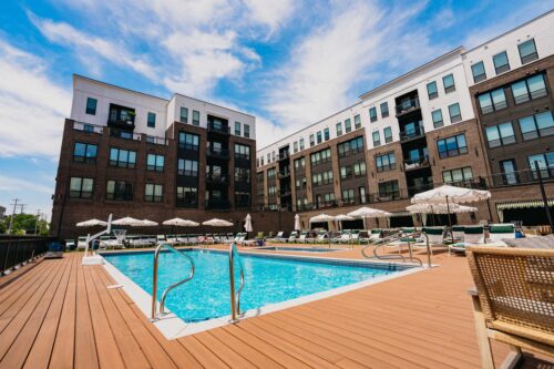 Outdoor swimming pool with lounge chairs and umbrellas, surrounded by a modern multi-story apartment building under a partly cloudy sky.