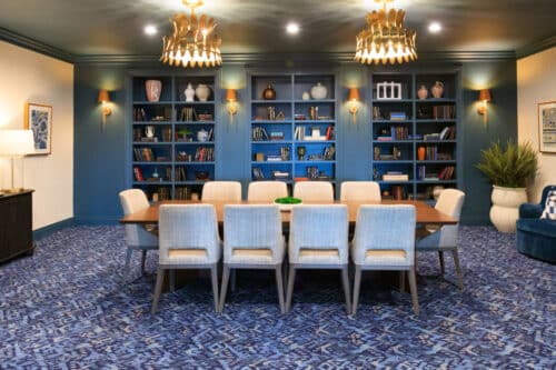 A modern conference room with a wooden table, ten upholstered chairs, blue bookshelves filled with decor and books, patterned carpet, and gold chandeliers.