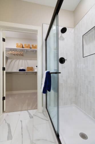 Modern bathroom with a glass-enclosed shower featuring white tile and black fixtures, opening to a walk-in closet with shelves, hangers, and storage baskets.