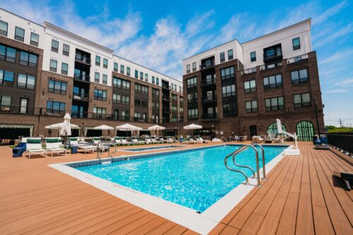 Rectangular outdoor swimming pool with poolside lounge chairs and umbrellas, surrounded by a large wooden deck and multi-story apartment buildings in the background.